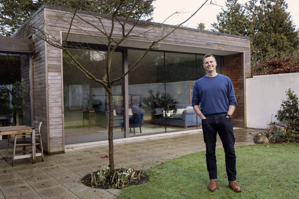 Paul Moore, Director of McCann Moore Architects, stands in the garden of a modern cedar-clad house